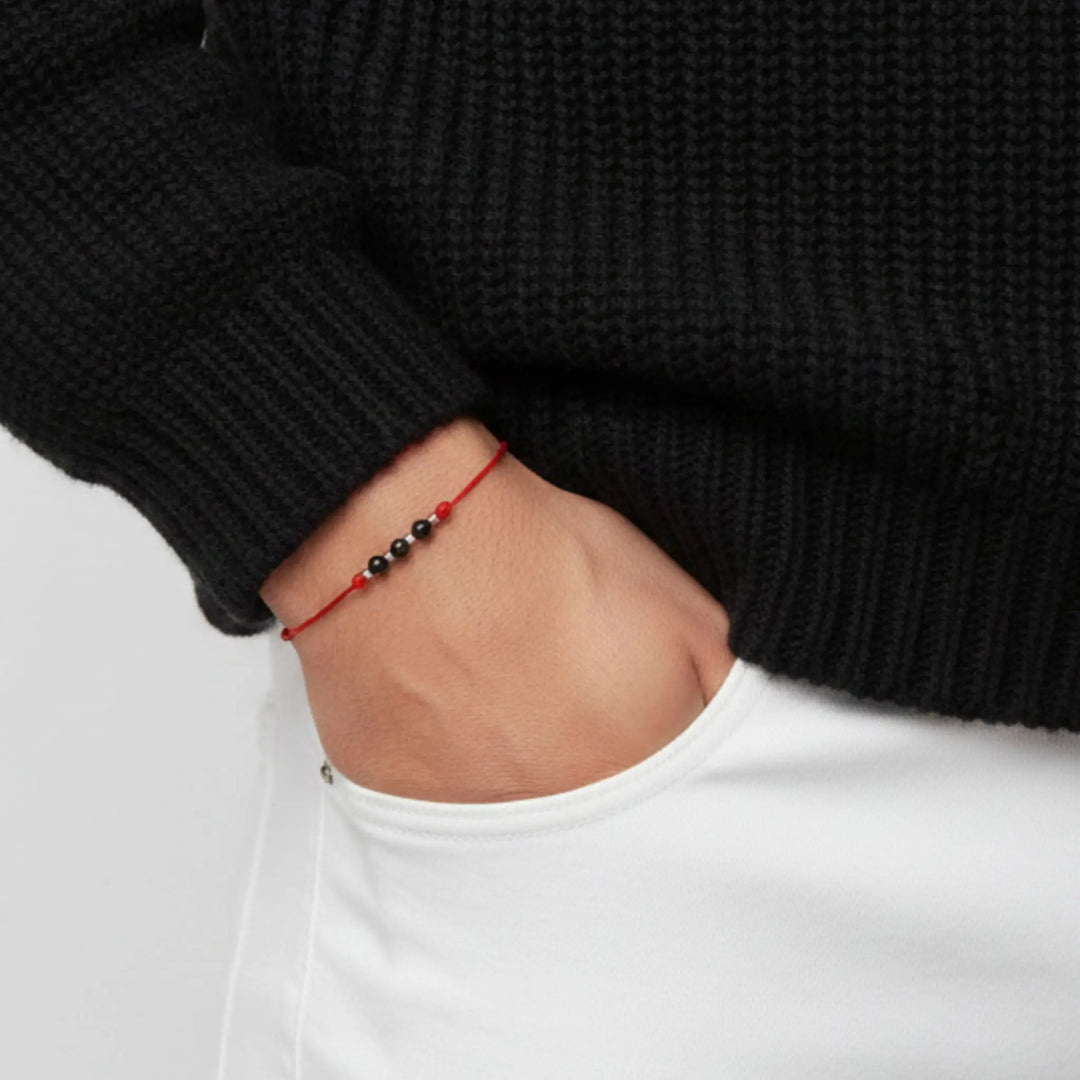 Person wearing a black sweater and white pant with a red and black tourmaline beaded bracelet on a plain background