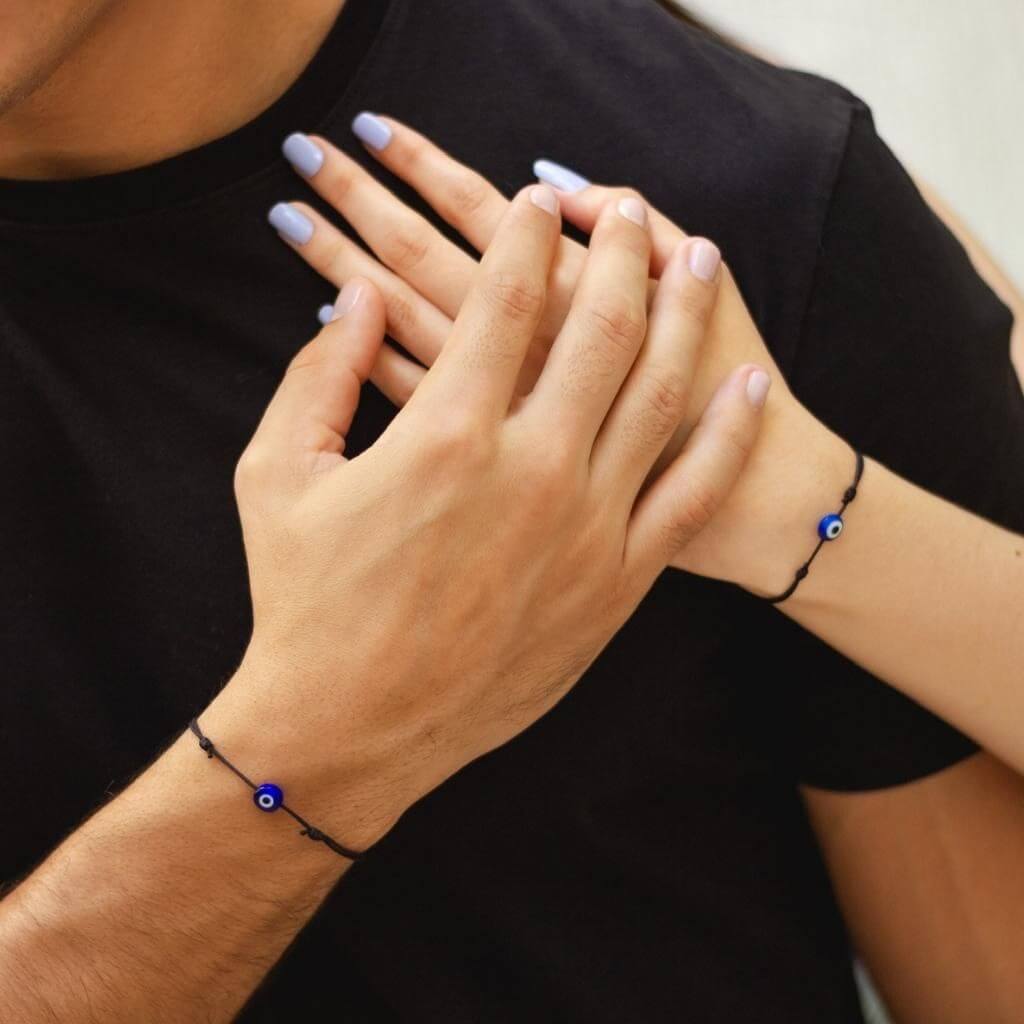 Hands together showcasing black string bracelets with evil eye charms, unisex style - Luck Strings.