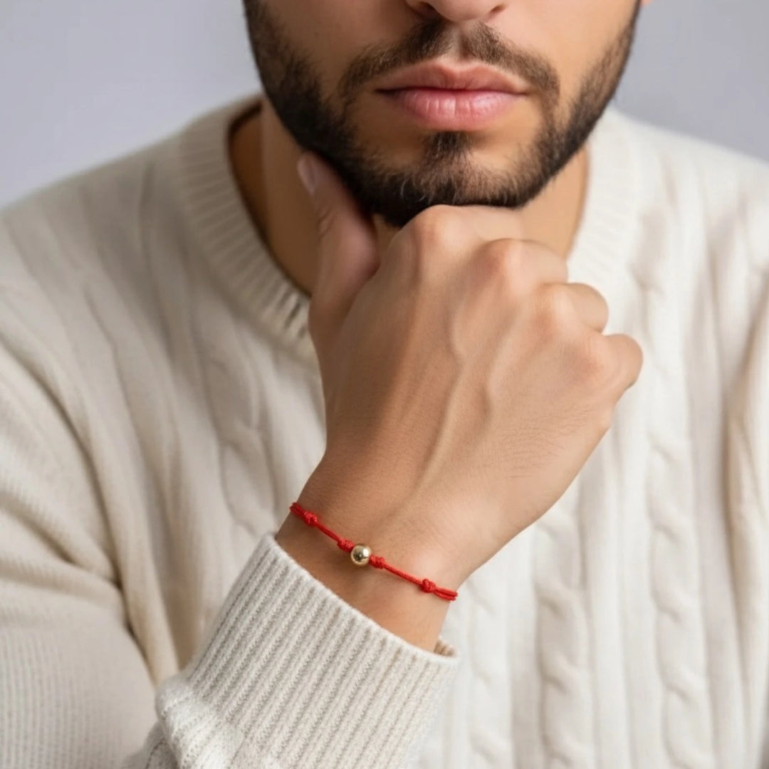 Man wearing a red bracelet with a small solid gold bead, wearing a beige sweater.