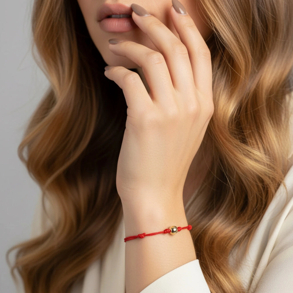 Close-up of a woman's hand with a red solid gold bead bracelet, wearing a white outfit against a neutral background
