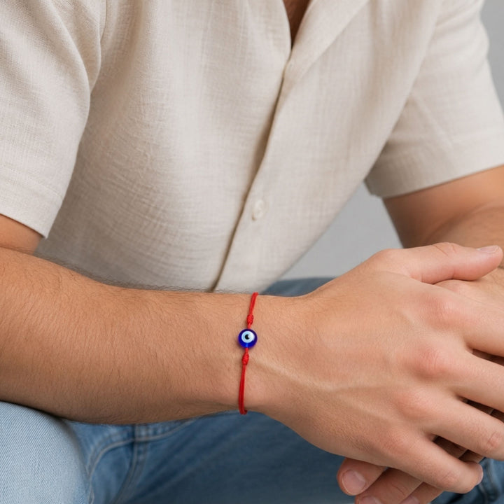 A close-up of a person's wrist wearing a red cord bracelet with a blue and white evil eye charm, paired with a light beige short-sleeve shirt and blue jeans.