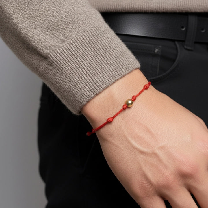 Hand wearing a red string bracelet with a gold bead, against a neutral background.