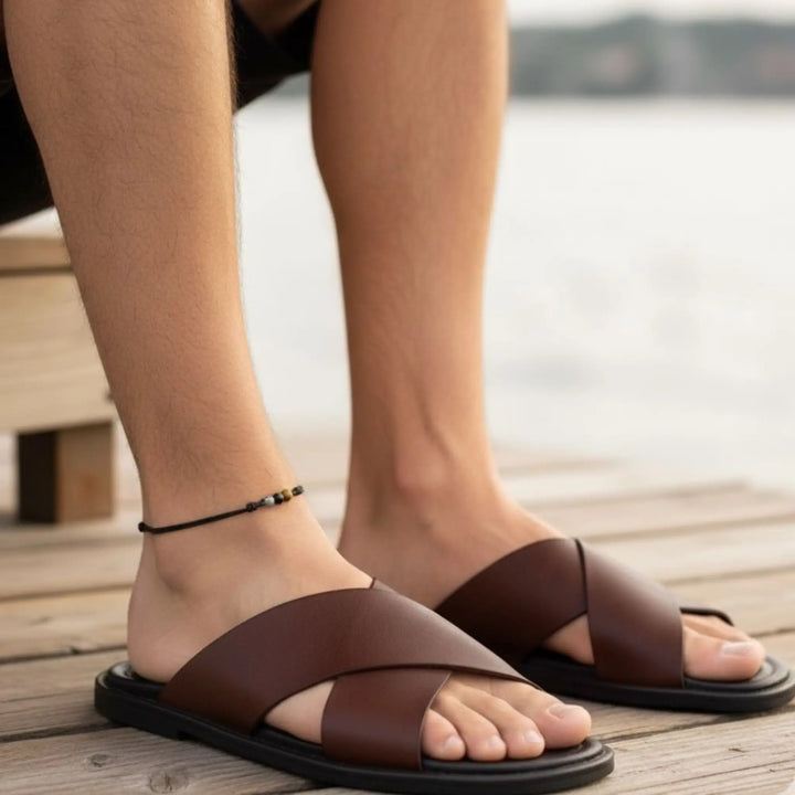 Brown sandals worn worn with a black anklet on feet standing on a wooden deck with a blurred background 