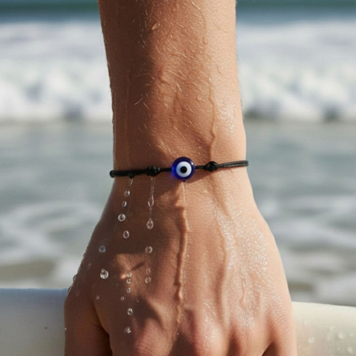 Hand wearing a black bracelet with a blue evil eye bead by the sea.