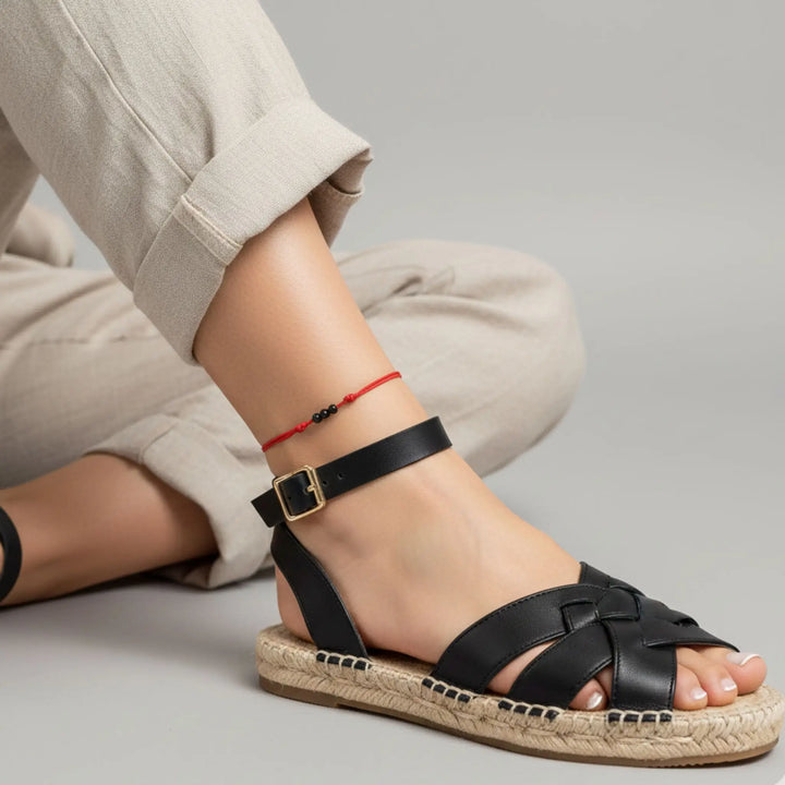 Red string anklet with three natural black tourmaline beads on a woman’s ankle, styled with black sandal with a gold buckle worn by a person on a gray background