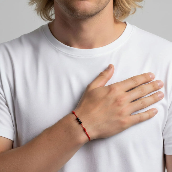 Person wearing a white t-shirt with a hand resting on their chest, wearing a red string black tourmaline bracelet.