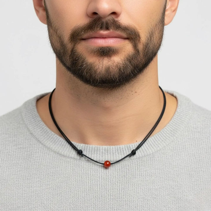 Man wearing a black carnelian necklace with a red bead on a plain background