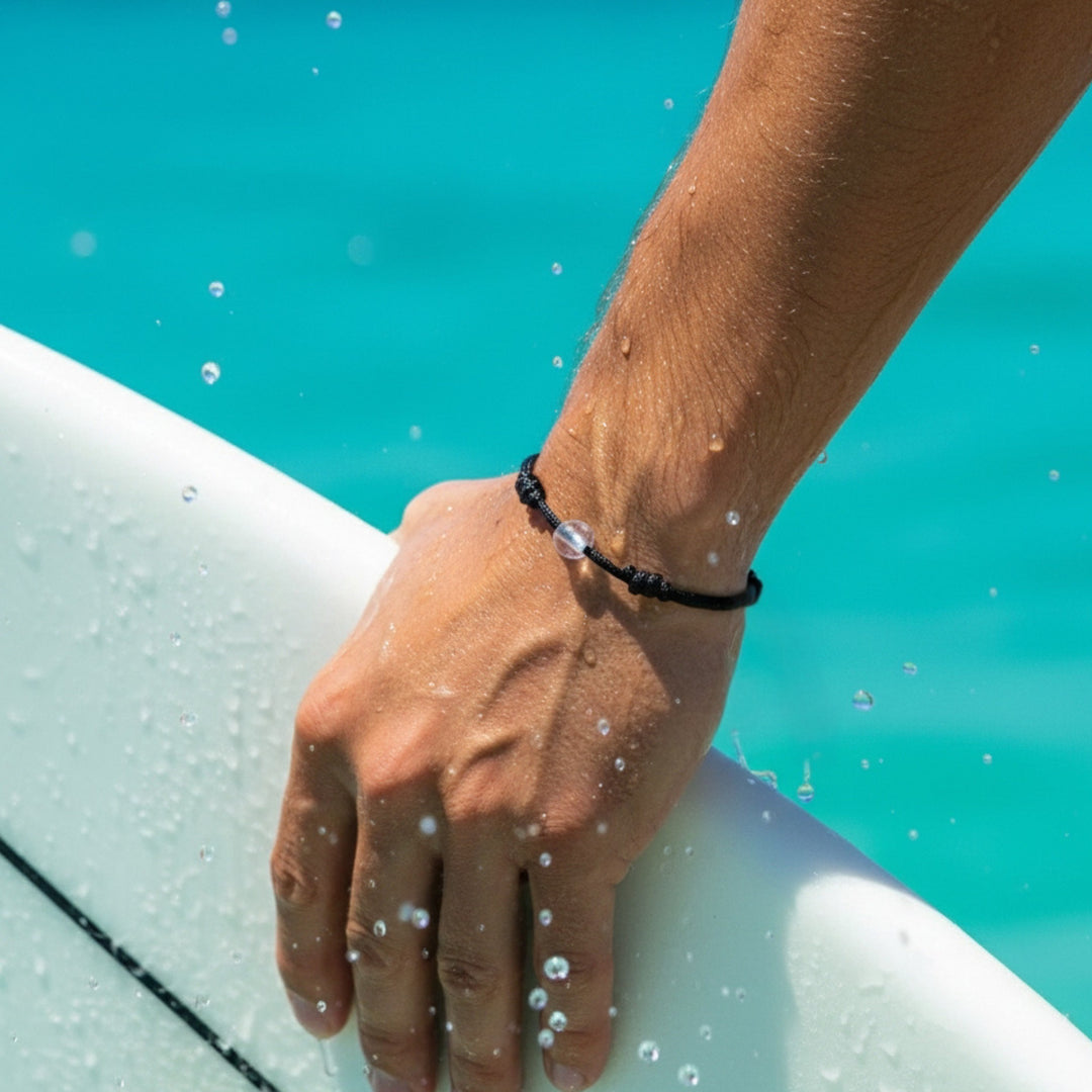 Man wearing a black cord bracelet with crystal quartz beads on his wrist while holding a surfboard.
