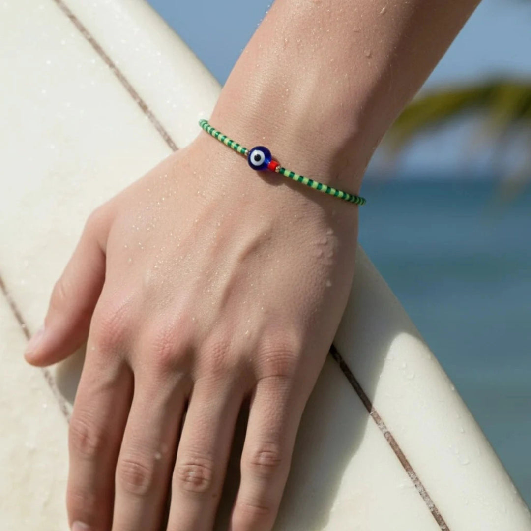 Hand wearing a green bracelet with a blue eye design on a surfboard by the ocean.