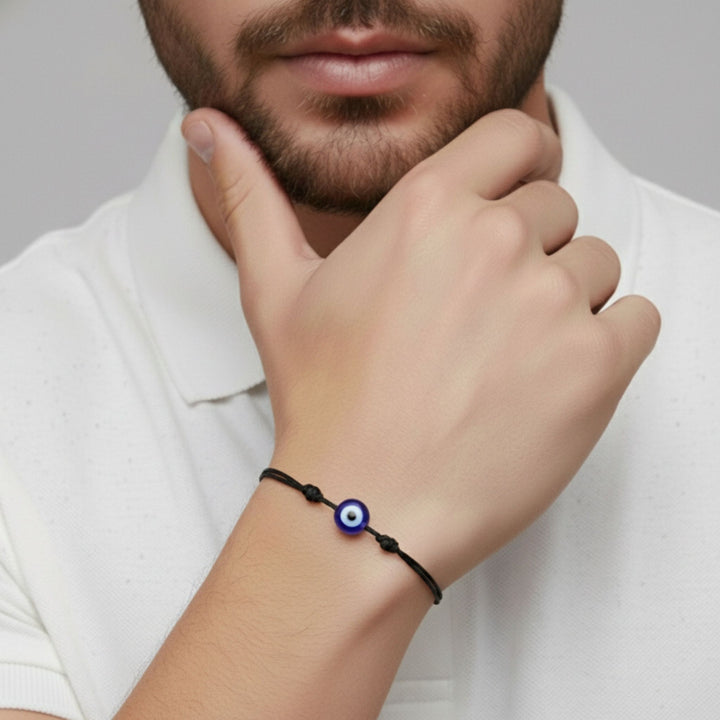 A close-up of a man's hand and wrist wearing a black cord bracelet with a blue and white evil eye charm, paired with a white polo shirt against a neutral gray background.