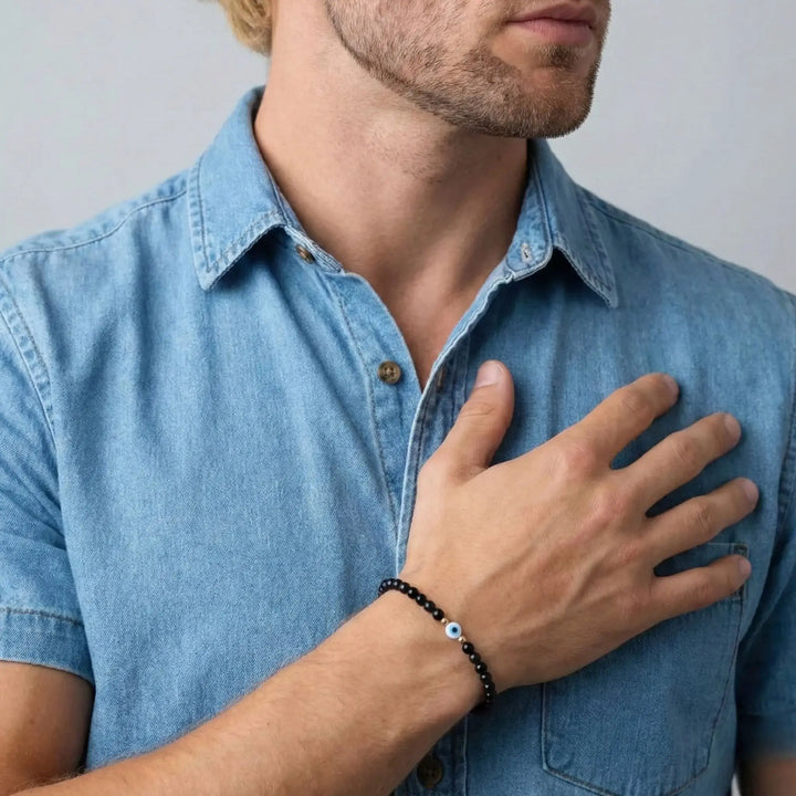 Man wearing a blue denim shirt with a evil eye black tourmaline bracelet on his wrist, touching his chest.