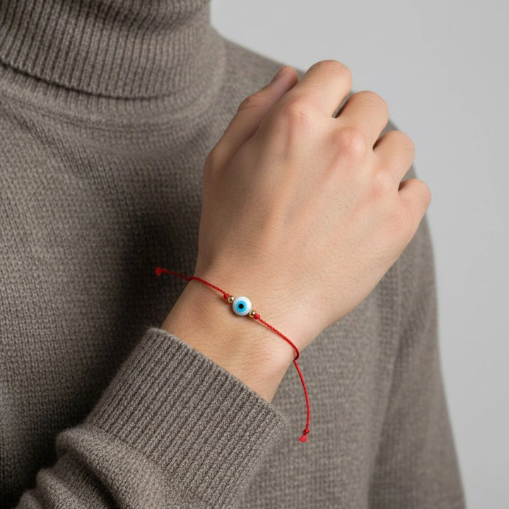 Hand wearing a red string bracelet with a blue bead, against a neutral background.
