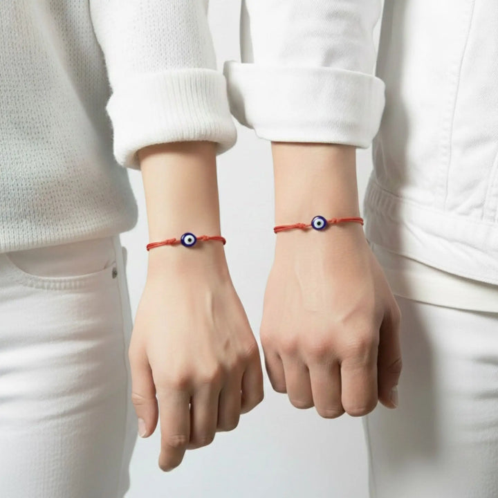 Two hands wearing red bracelets with blue evil eye symbols on a plain background