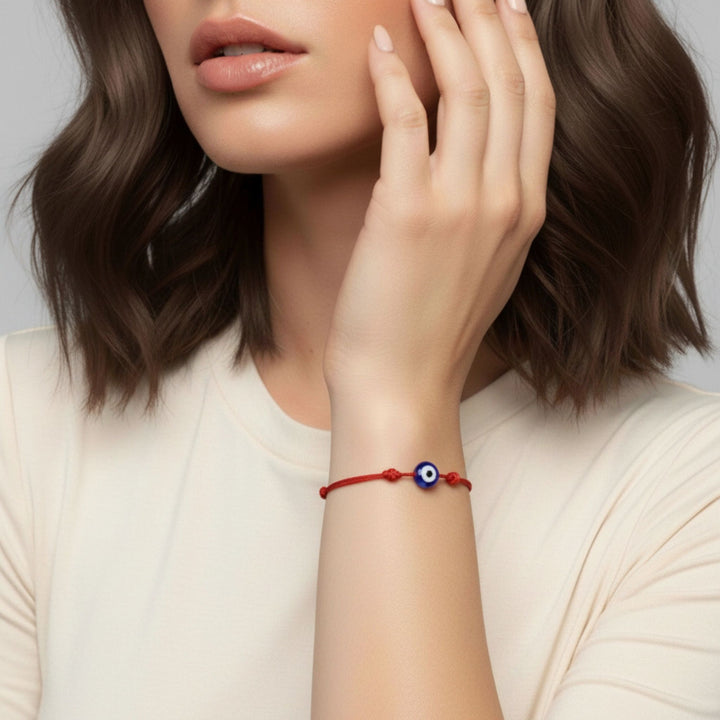 A close-up of a woman wearing a red cord bracelet with a blue and white evil eye charm on her wrist, paired with a cream-colored top.