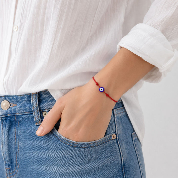 A close-up of a person wearing a red cord bracelet with a blue and white evil eye charm, styled with a white button-up shirt and blue jeans.