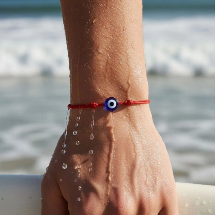 Hand with a red string bracelet featuring an evil eye charm against a beach background