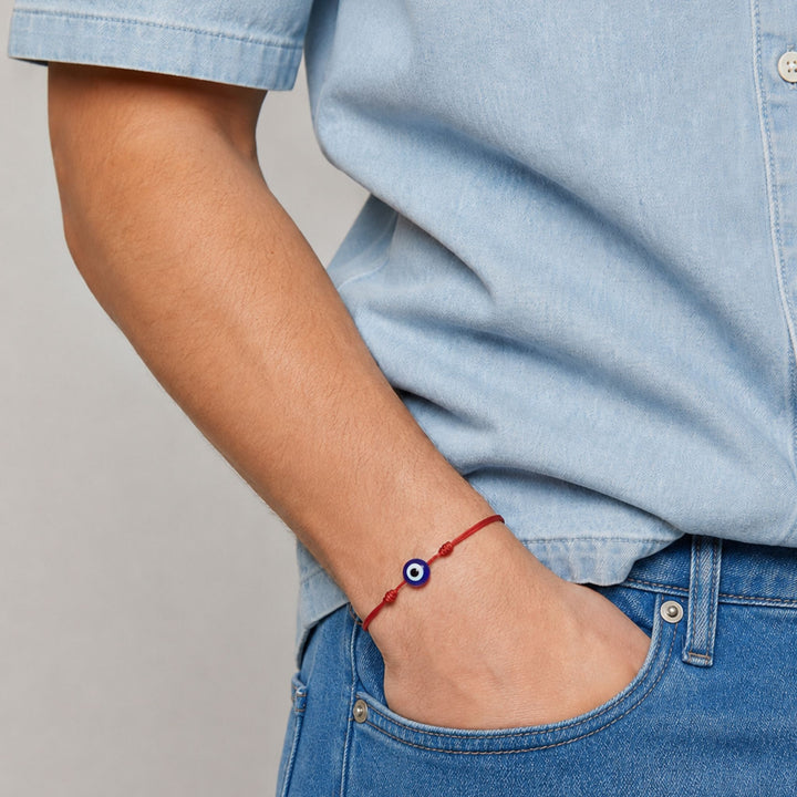 A close-up of a person's wrist wearing a red cord bracelet with a blue and white evil eye charm, paired with a light blue denim shirt and blue jeans.