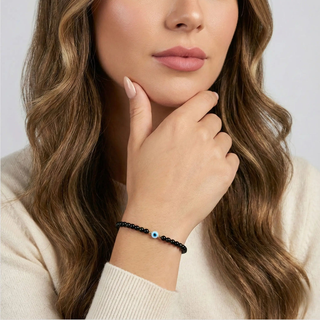 Woman wearing a black tourmaline beaded bracelet with a blue evil eye bead, touching her face against a neutral background