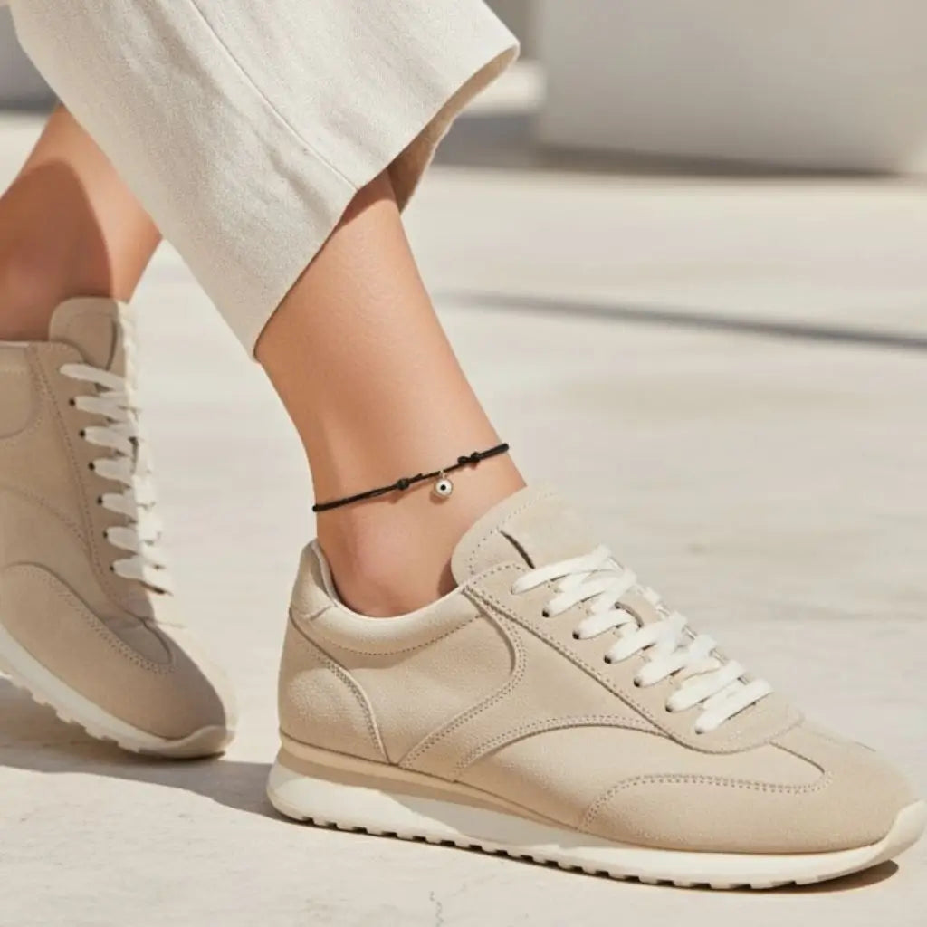 Beige sneakers worn with white pants and gold evil eye black anklet  worn by a woman on a blurred background