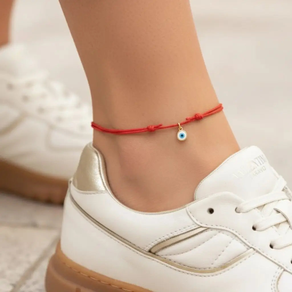 Red anklet with a gold blue evil eye charm on a person's ankle, wearing a white sneaker.