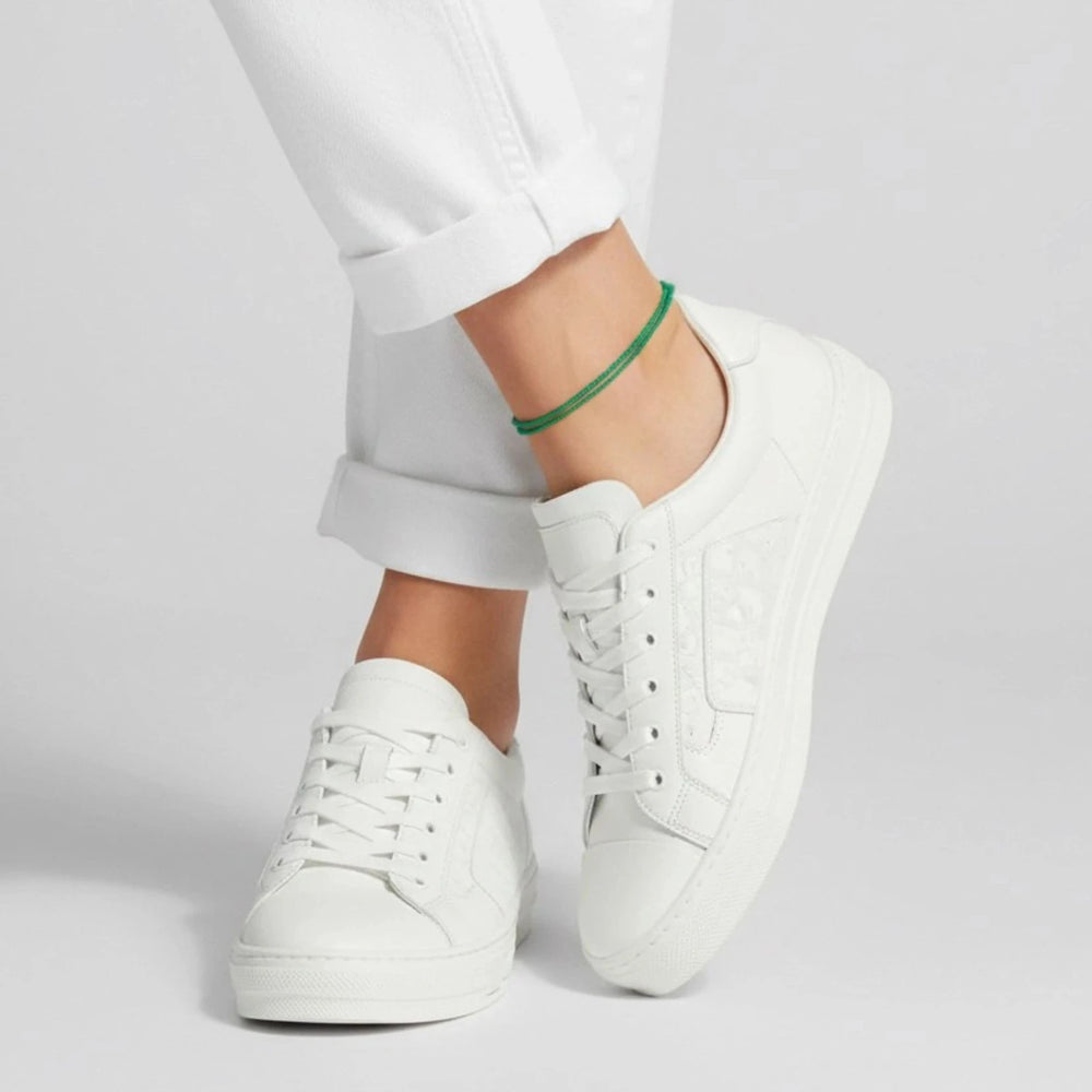 White sneakers worn with white pants and green cord anklet on a light gray background