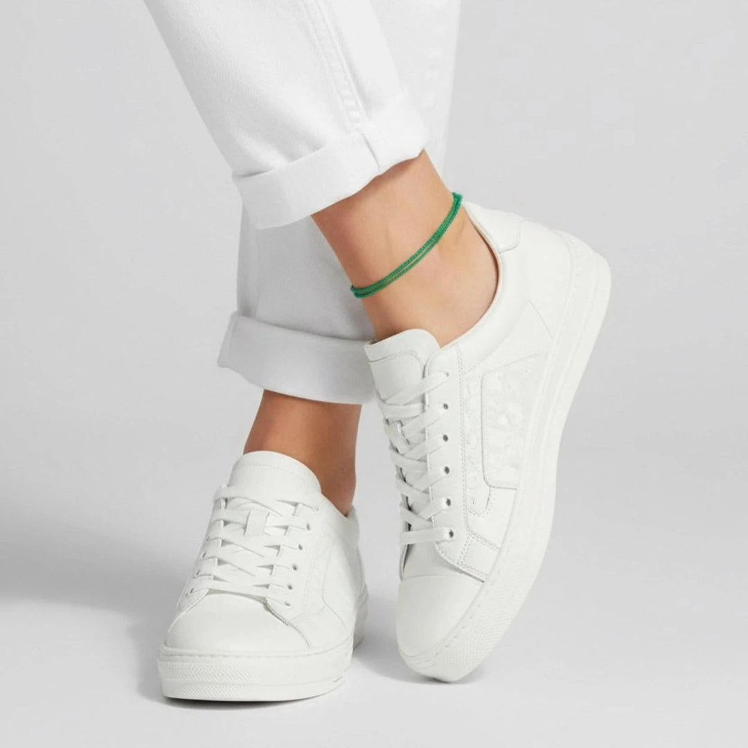 White sneakers worn with white pants and green cord anklet on a light gray background