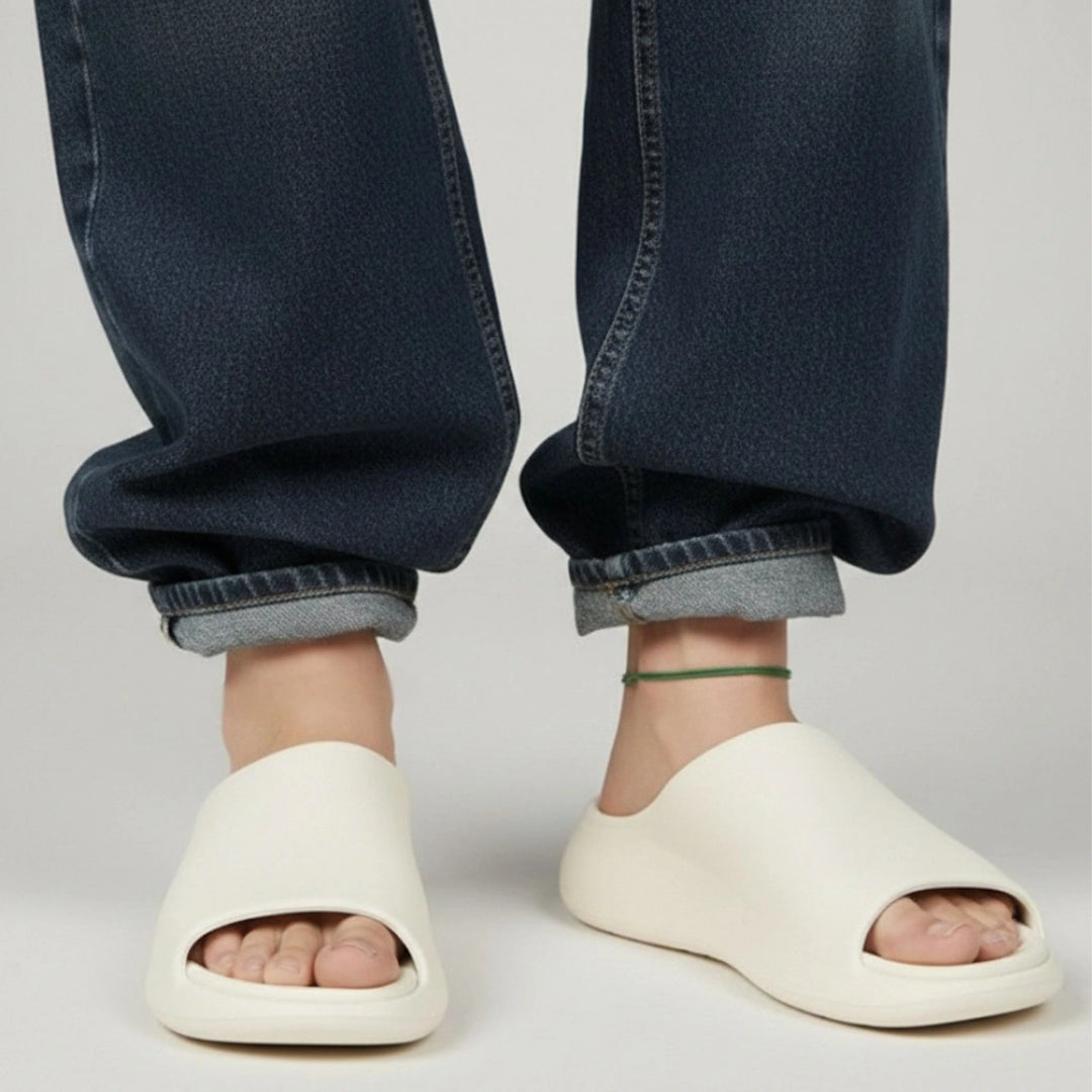 White open-toe sandals worn with rolled-up blue jeans and green cord anklet on a light gray background