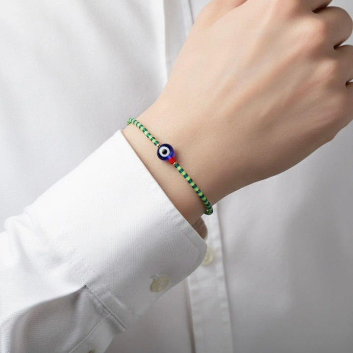 Hand wearing a green beaded bracelet with a blue and red eye charm on a white shirt.