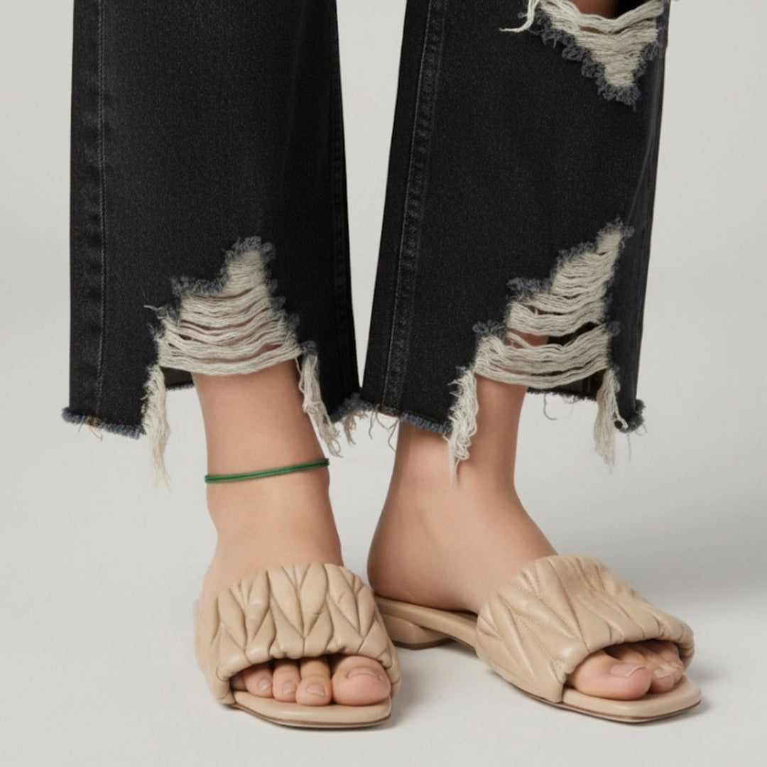 Beige open-toe sandals worn with ripped black jeans and green cord anklet on a neutral background
