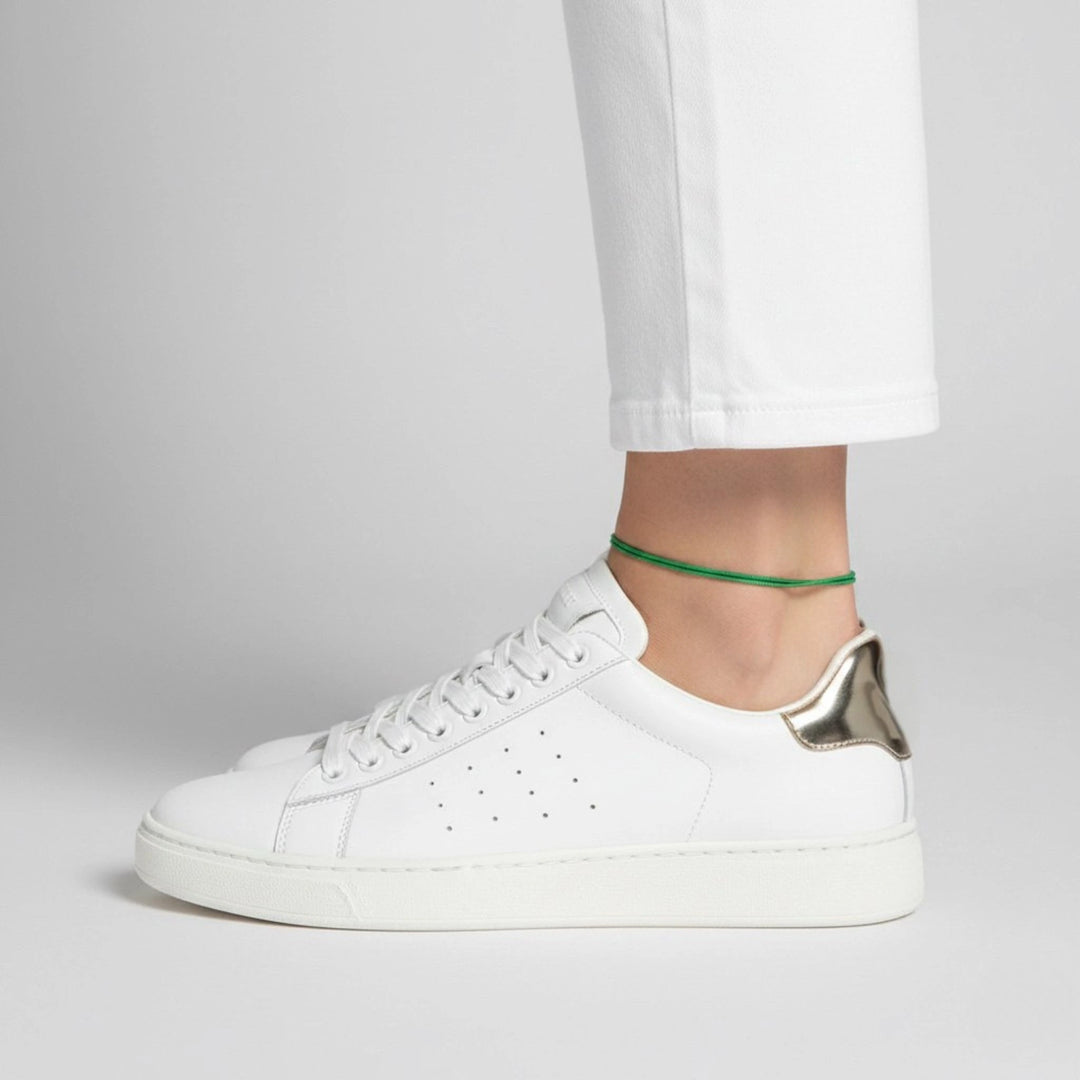 White sneaker worn with a plain background and green cord anklet 