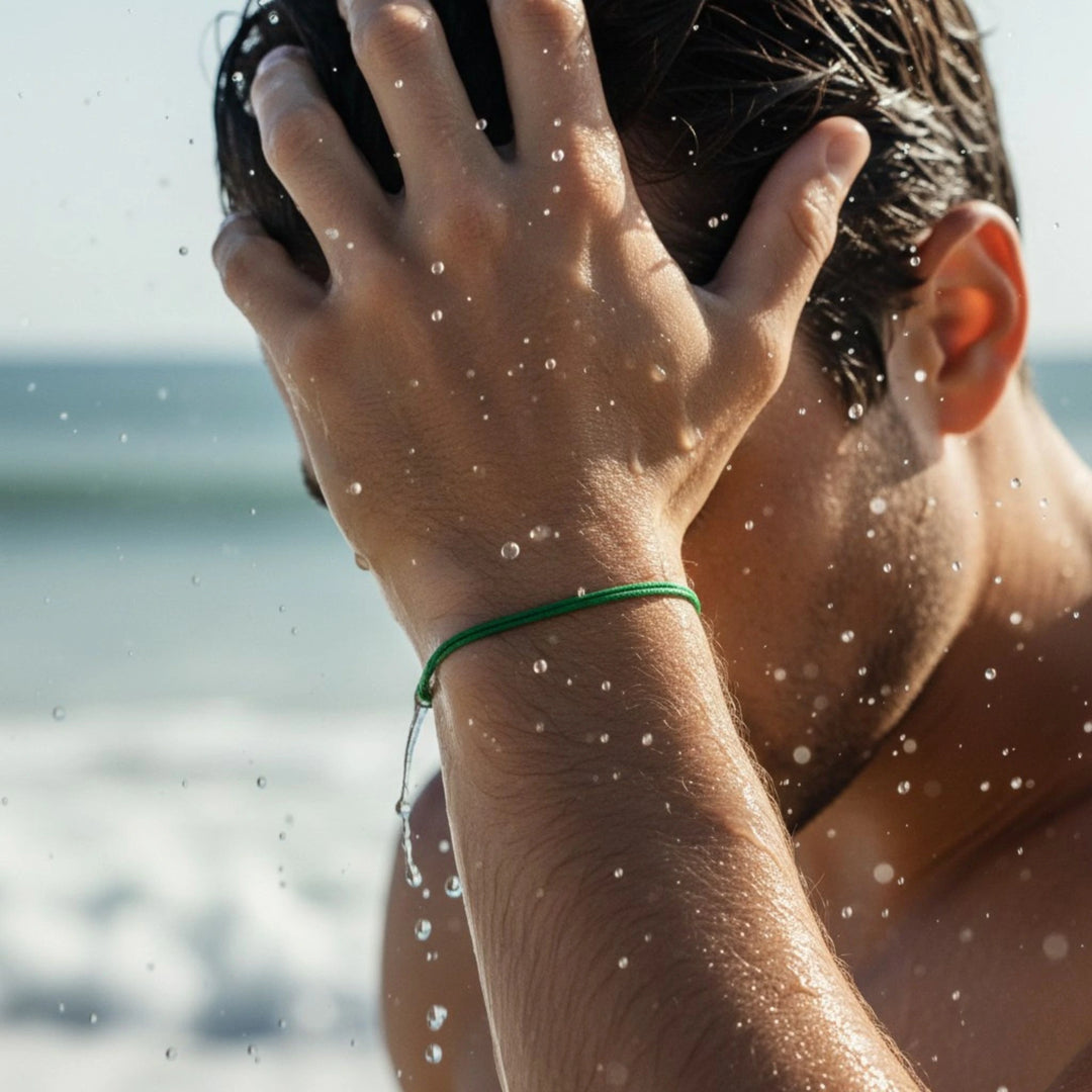Person with a green bracelet holding their head, with water droplets visible, against a blurred beach background.