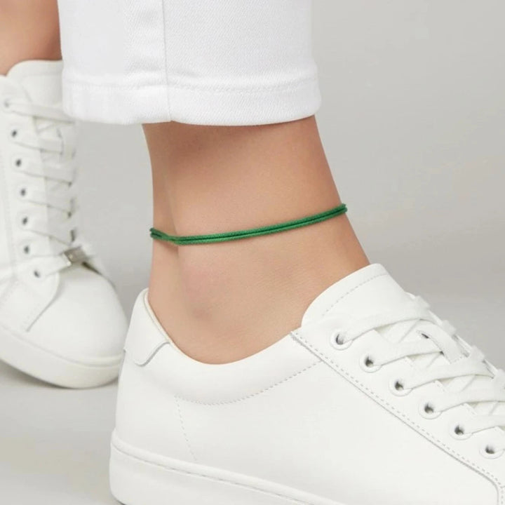 White sneakers worn with white pants and green cord anklet on a plain background