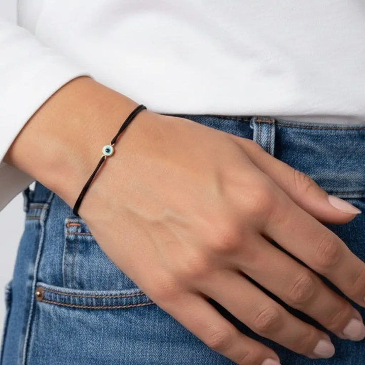 Hand wearing a black bracelet with a small gold evil eye connector bead, against a white shirt and blue jeans background.