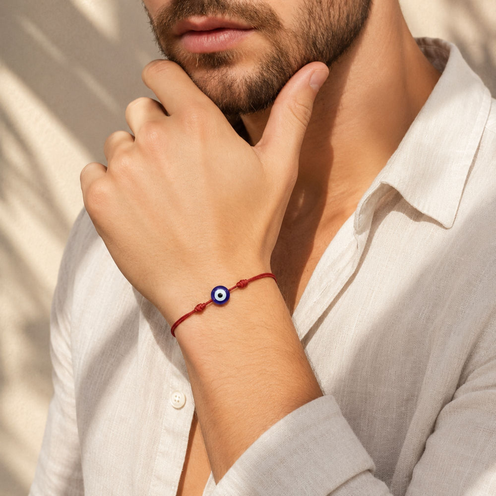 A close-up lifestyle shot of a man wearing a red cord bracelet with a blue and white evil eye charm, paired with a white linen shirt.