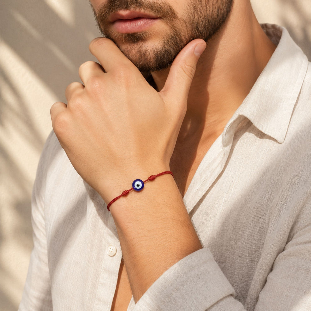 A close-up lifestyle shot of a man wearing a red cord bracelet with a blue and white evil eye charm, paired with a white linen shirt.