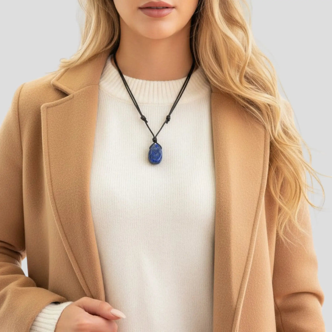 Person wearing a beige coat and white sweater with a blue lapis lazuli  pendant necklace on a light gray background