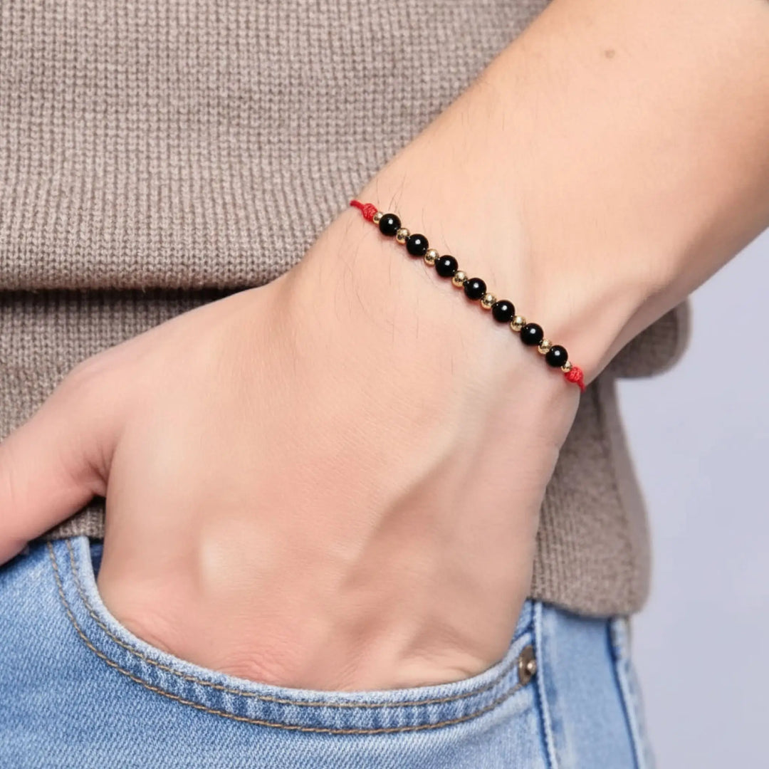 Hand wearing a beaded bracelet with black tourmaline red string and gold beads on a neutral background