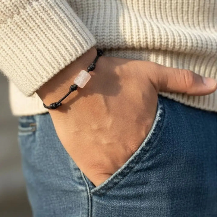 Hand wearing a bracelet with a rose quartz stone, wearing a beige sweater and blue jeans.