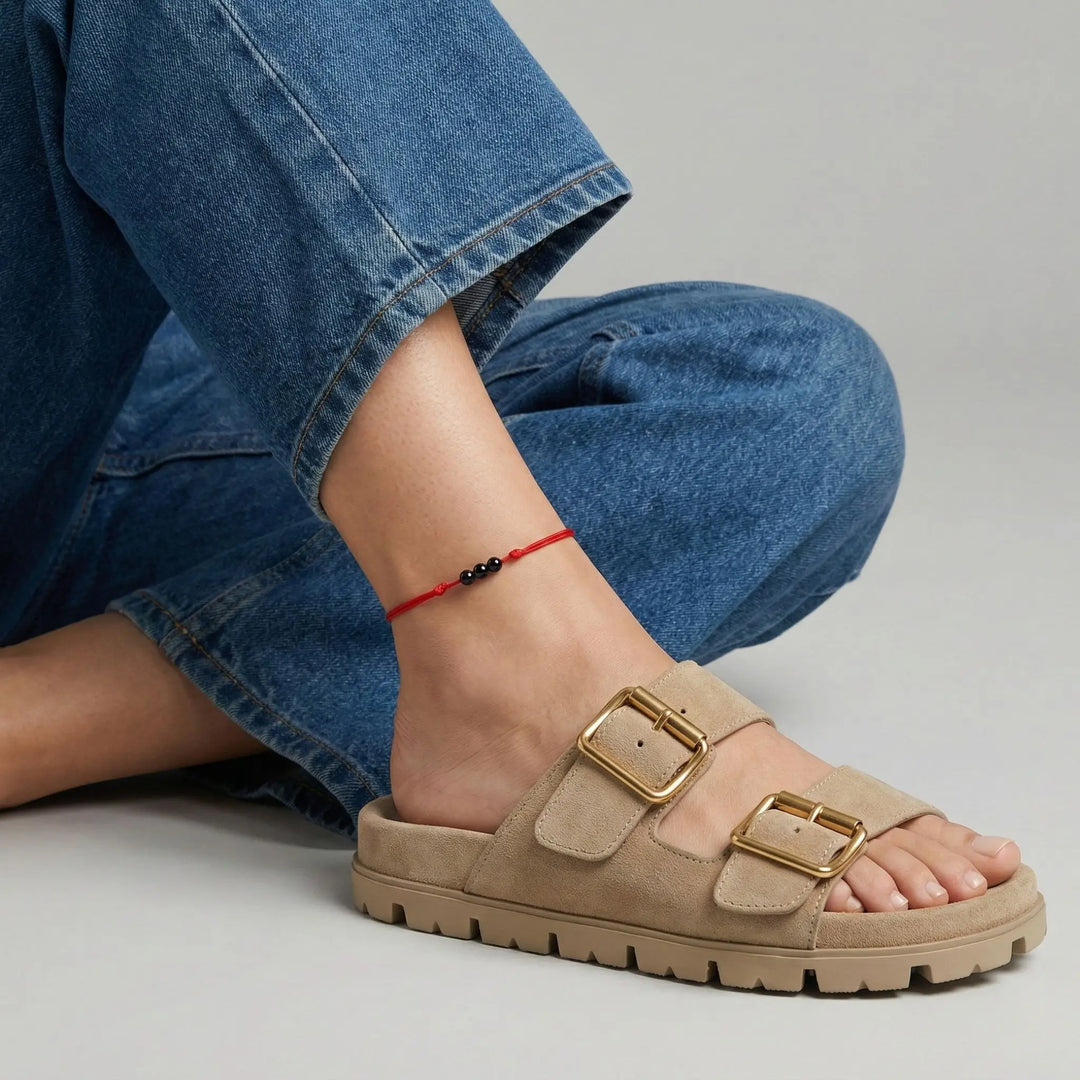 Red string anklet with three natural black tourmaline beads on a woman’s ankle, styled with beige sandals with gold buckles worn with blue jeans on a neutral background