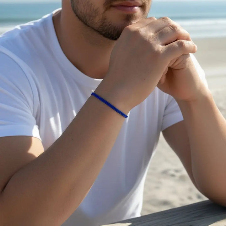 Man sitting on a beach with hands clasped, wearing a blue bracelet.