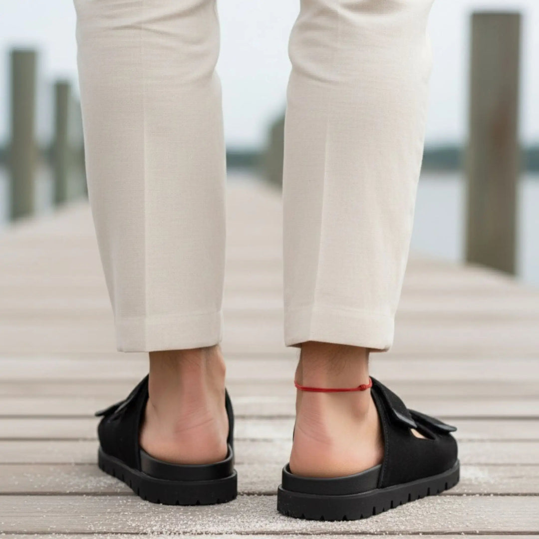 Men’s red string anklet styled with black flat shoes worn with beige pants on a wooden deck.  