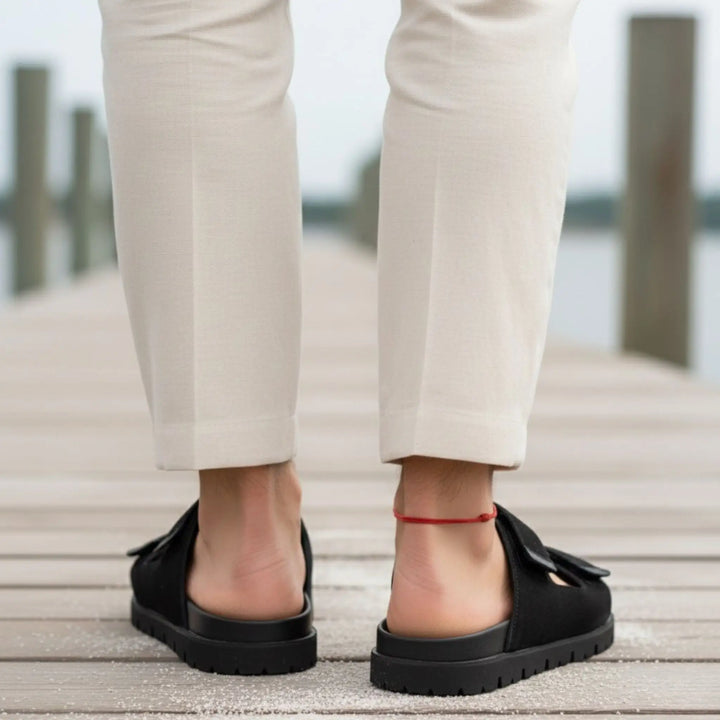 Men’s red string anklet styled with black flat shoes worn with beige pants on a wooden deck.  