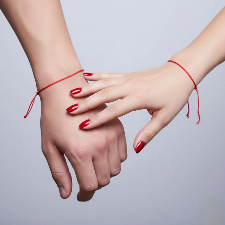 Two hands holding each other with red nail polish and red strings against a light gray background