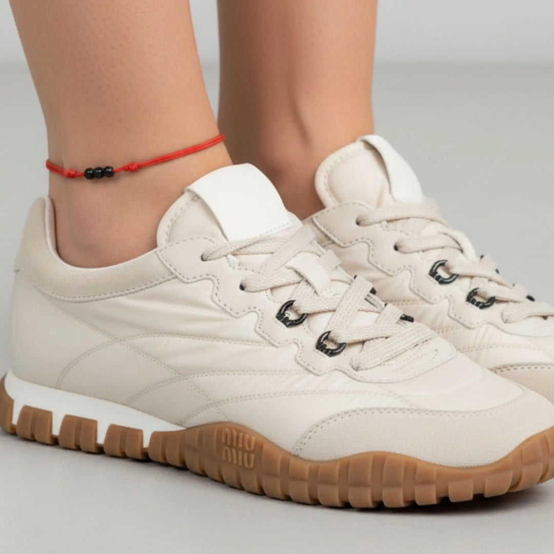 Black string anklet with three natural black tourmaline beads on a woman’s ankle, styled with beige sneakers with brown soles worn by a person on a light gray background