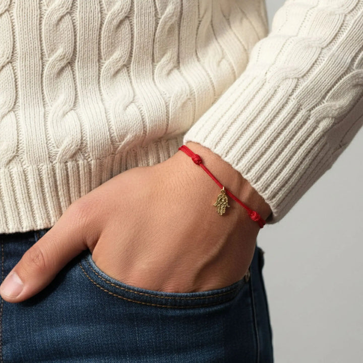 Hand wearing a red gold hamsa bracelet with a gold charm, against a neutral background