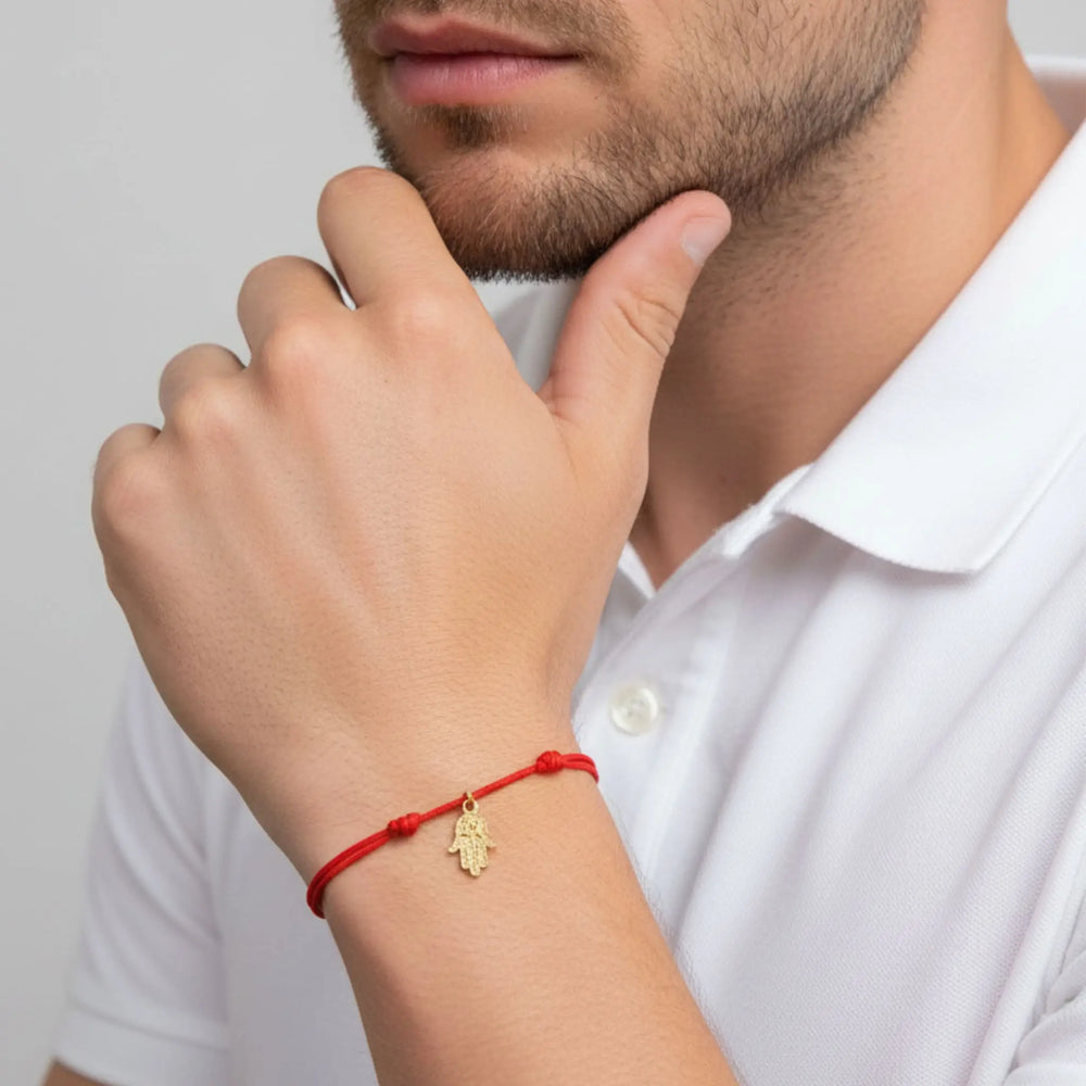 Person wearing a red gold hamsa bracelet with a gold charm on a plain background
