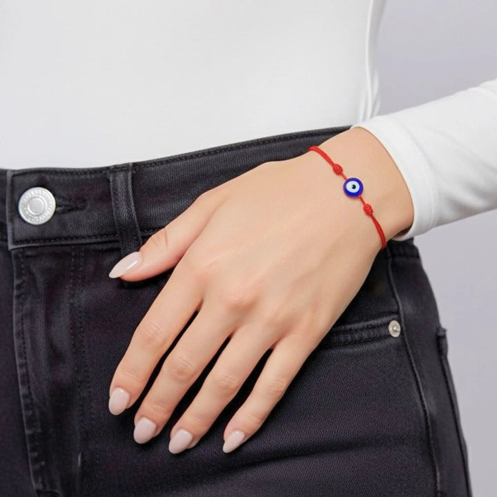 Hand wearing a red bracelet with a blue evil eye charm on a plain background