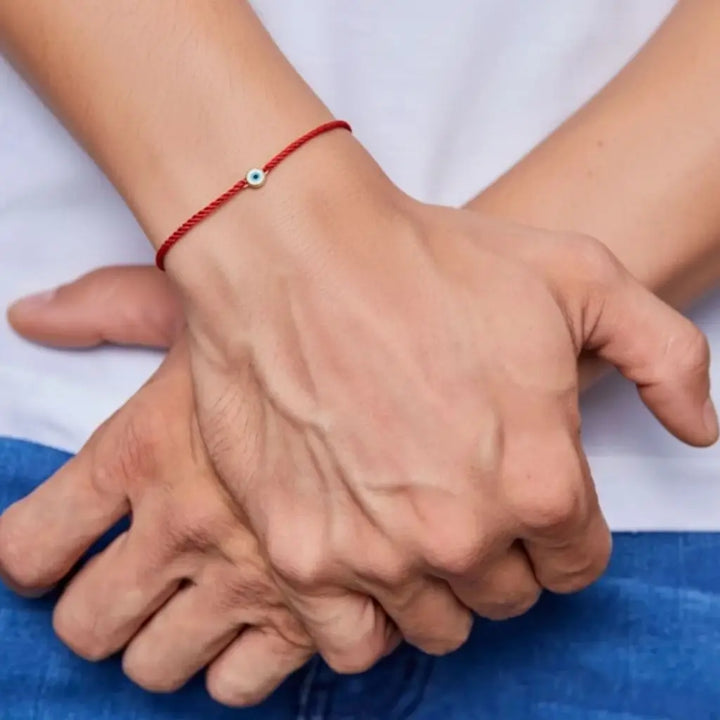 Two hands holding each other with a red evil eye string bracelet on a blue and white background
