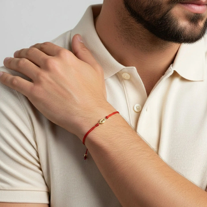 Person wearing a gold evil eye red bracelet with a heart charm on a plain background