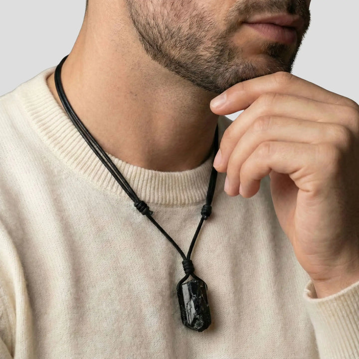 Man wearing a beige sweater with a black necklace, touching his chin.